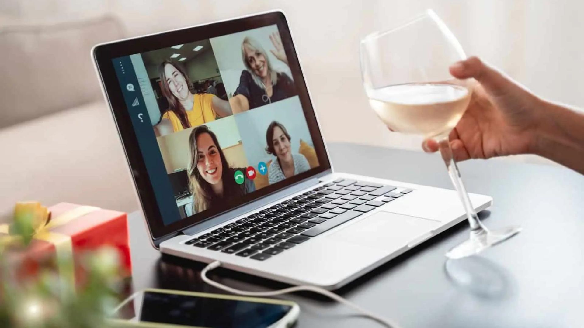 a hand holding a glass of wine and a laptop in the front following virtual partying 